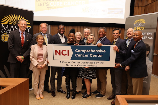 Massey leaders and elected officials stand in front of an auditorium posing with an NCI Comprehensive Cancer Center badge