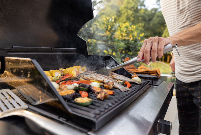 Person flips vegetables and fish with tongs on a grill